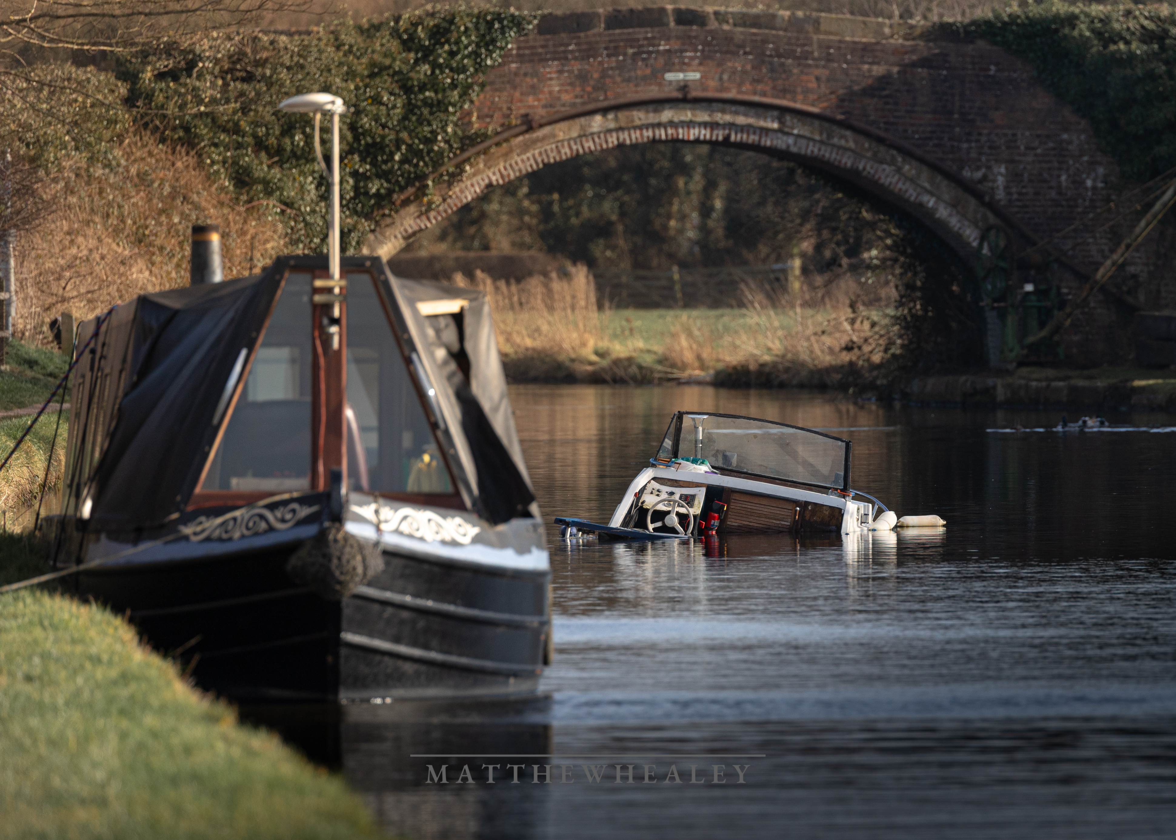 Sinking boat on English canal beside narrowboat with brick arch bridge in background.